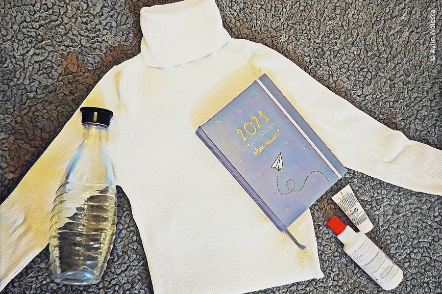 Eine leere Glasflasche, ein Rollkragenpullover, ein Kalender und Cremes liegen auf einem Teppichboden.